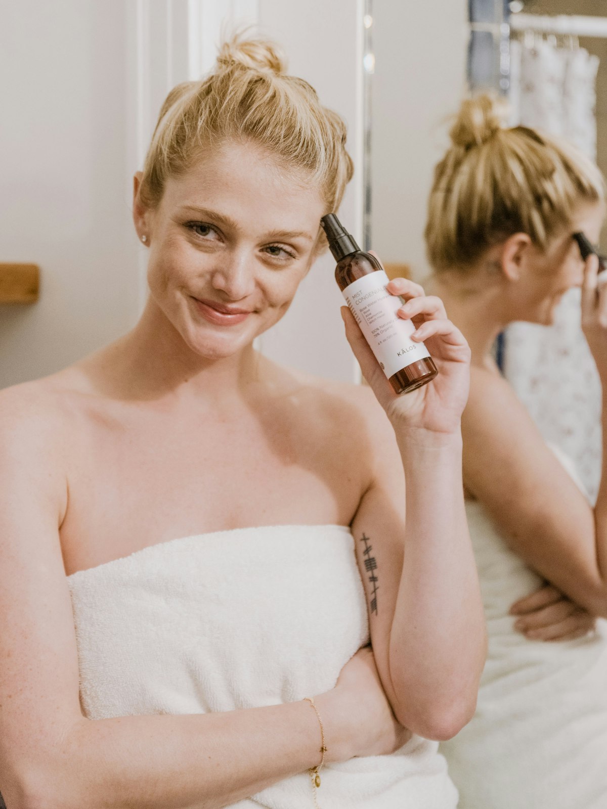Woman applying body oil to her shoulder after a shower, wrapped in a towel in steamy bathroom light