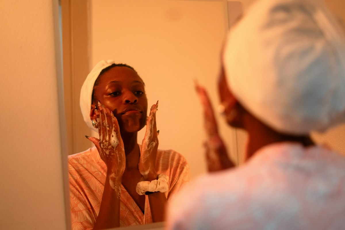 Woman removing her makeup with a cotton pad at the bathroom sink in the evening, towels and gentle cleanser beside her
