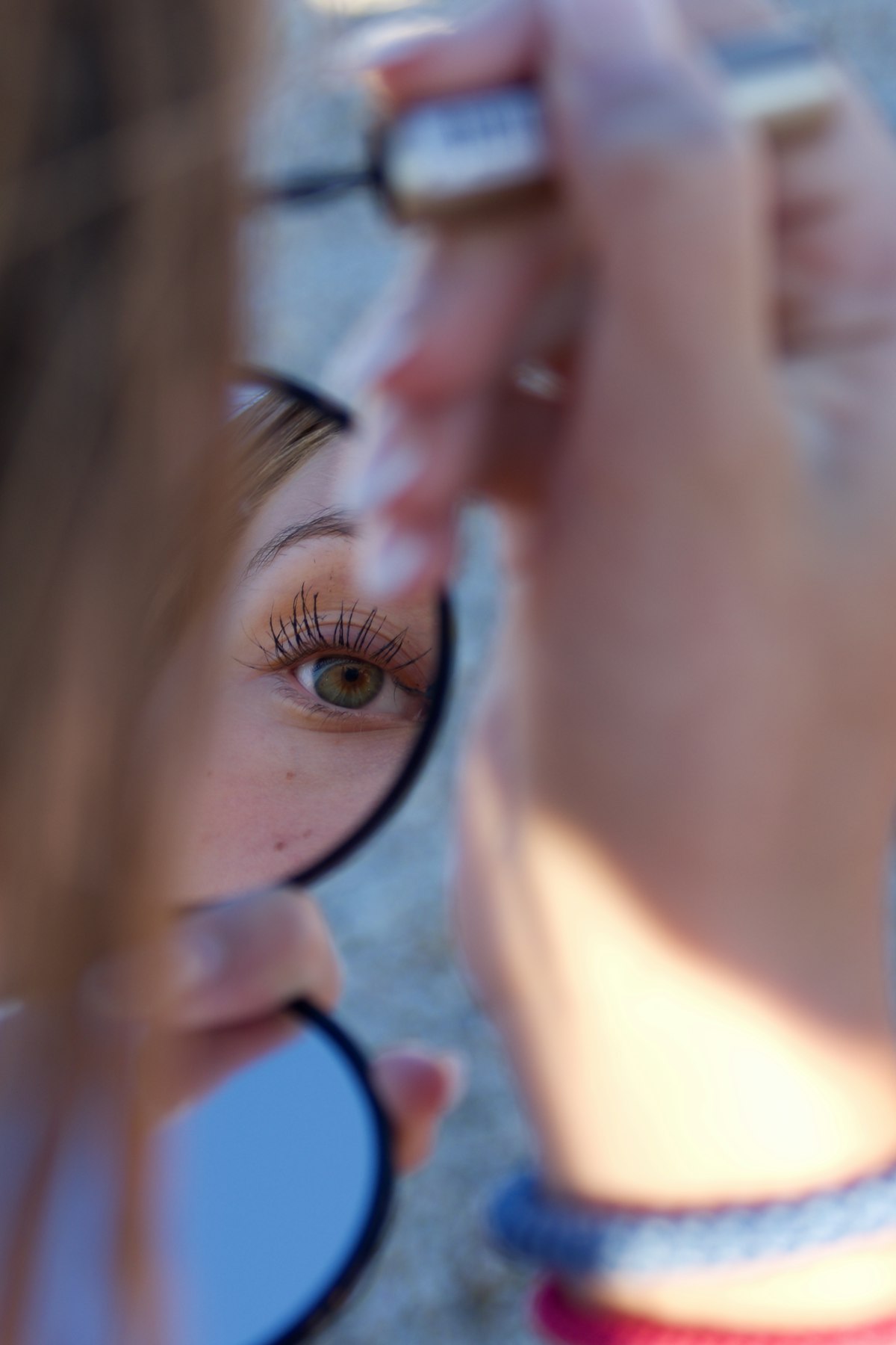 Woman applying mascara to her upper lashes while getting ready in front of a bedroom mirror